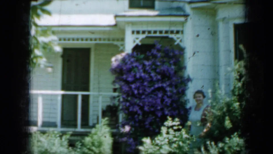 1958 Minnesota 8mm frame showing a white clapboard porch, dense purple flowers, garden greenery, and a woman at frame right.