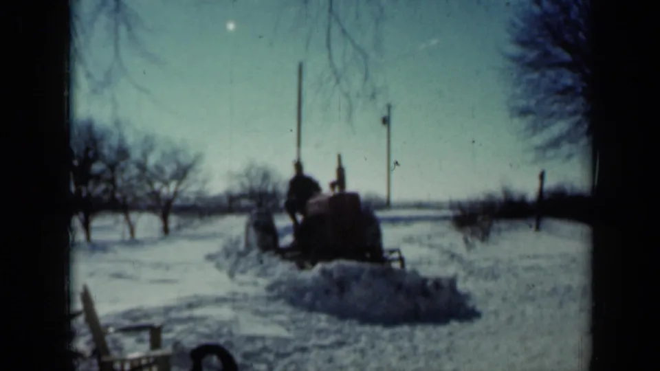 A man drives a tractor fitted with a makeshift plow through deep snow in a 1958 Minnesota farmyard.