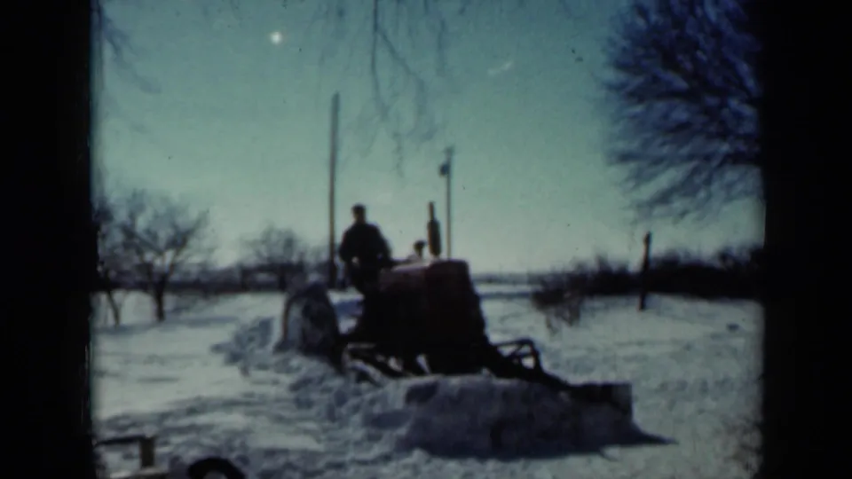 A distant man operates a tractor-mounted blade to push snow across a rural Minnesota farmyard in 1958.