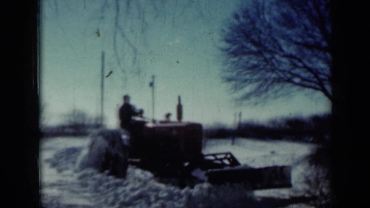 In 1958 Minnesota home-movie footage, a man drives a vintage tractor with a plow through snow near bare trees and utility poles.