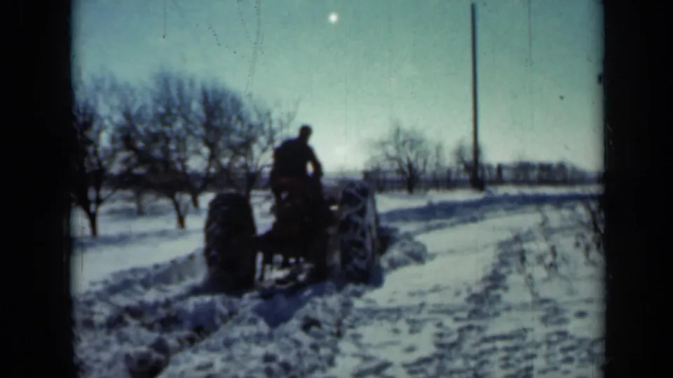 A man on a tractor drives away down a snow-covered rural road, framed by winter trees and a tall utility pole.