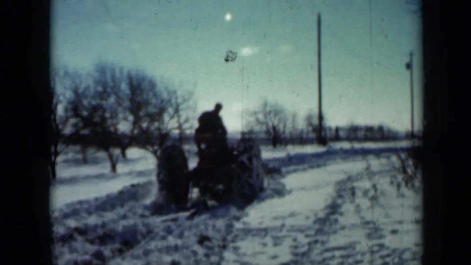 The tractor continues down the rural Minnesota road, with snow tracks, bare trees, and utility poles visible under a pale winter sky.