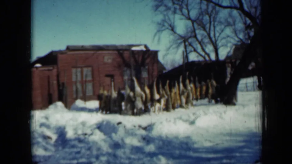 1958 Minnesota home-movie view of corn stalks lined in snow beside a red brick outbuilding, with bare trees overhead.