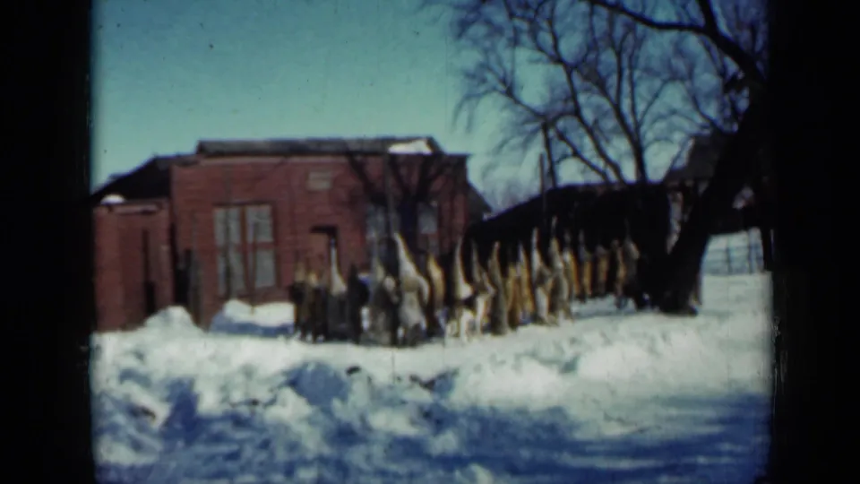 1958 Minnesota winter frame of upright corn stalks, snowbanks, a red brick building, and leafless trees in a rural yard.