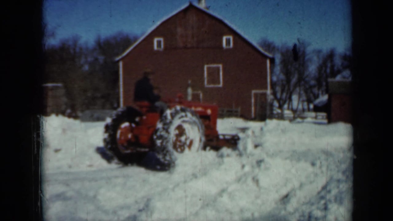 Red tractor with a seated operator plowing snow in front of a red barn.
