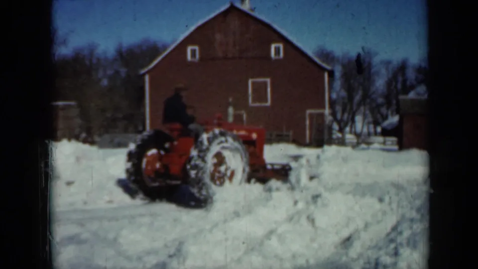 1958 Minnesota winter farm scene with a red tractor and seated operator clearing snow beside a large red barn with white-trimmed openings.