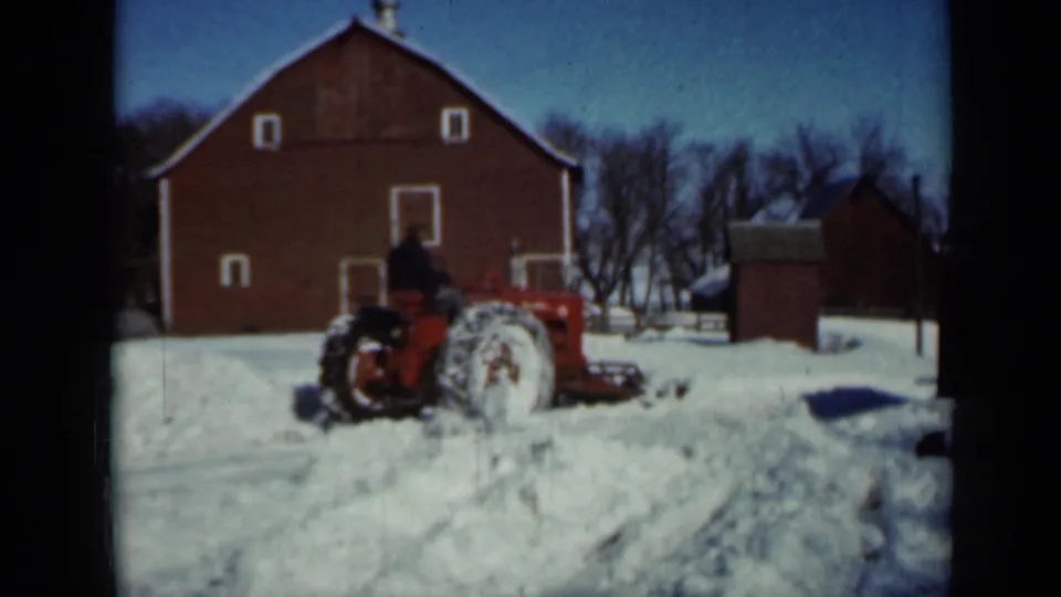 1958 Minnesota farm footage showing a red tractor with an operator pushing through deep snow near a red barn and a small outbuilding.