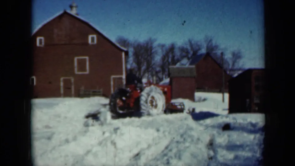 1958 Minnesota winter farmyard with a red tractor and operator clearing snow between a large red barn, smaller red outbuilding, and bare trees.