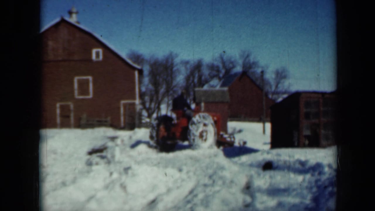 Red tractor clearing snow near a large red barn and small farm buildings.