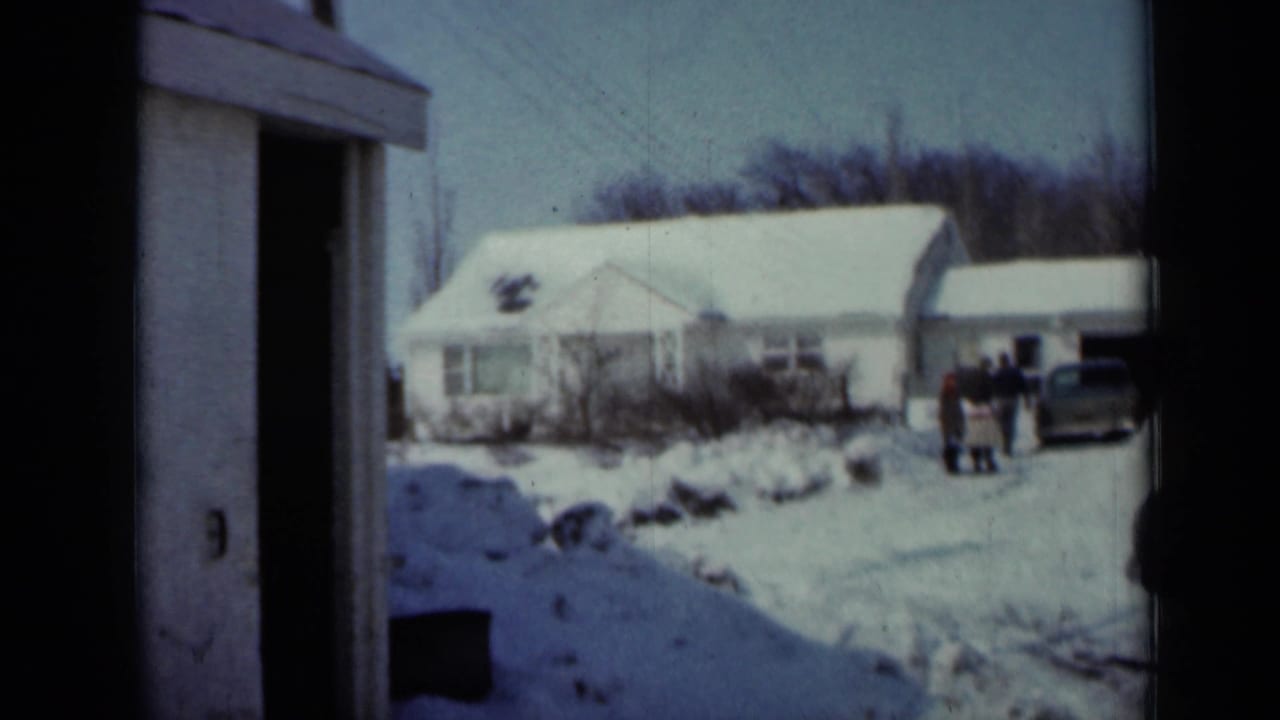 Snowy farmhouse yard with people near a car and a building edge in foreground.