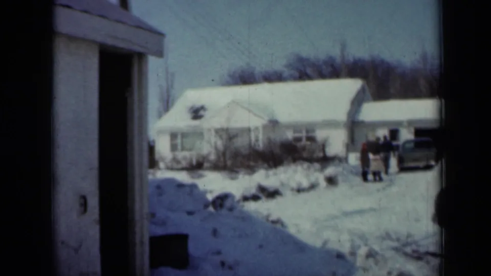 1958 Minnesota home-movie frame showing a snow-covered farmhouse yard, distant people, a parked car, and a dark building edge.