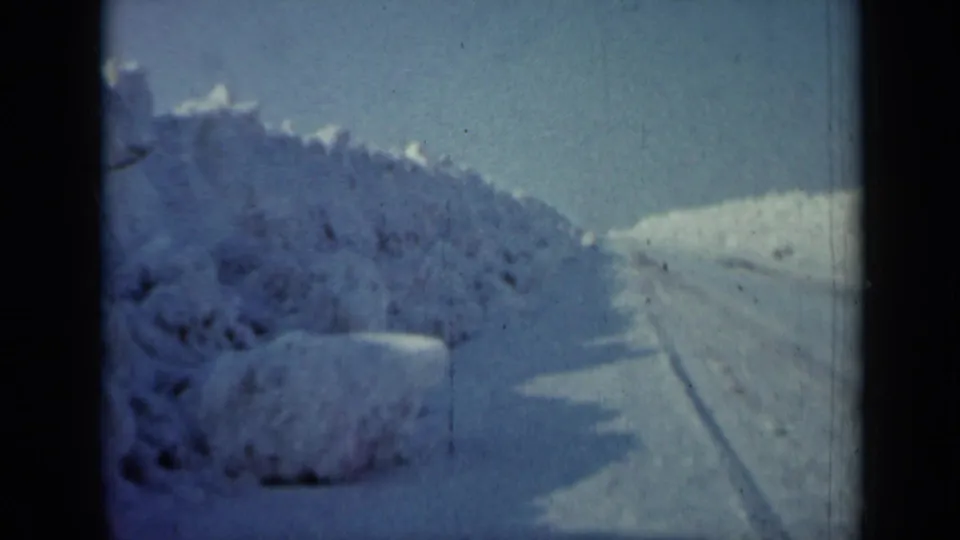 1958 Minnesota home-movie frame showing a plowed snowy road with deep snowbanks and dark brush along the left side.