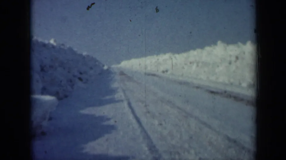 Archival 8mm frame from Minnesota in 1958 showing a quiet snow-covered road bordered by heavy drifts.