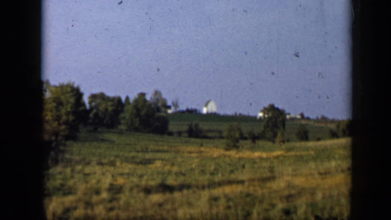 1959 Aledo, Texas 8mm frame showing open grassland, trees, and a distant white house or barn on a low hill.