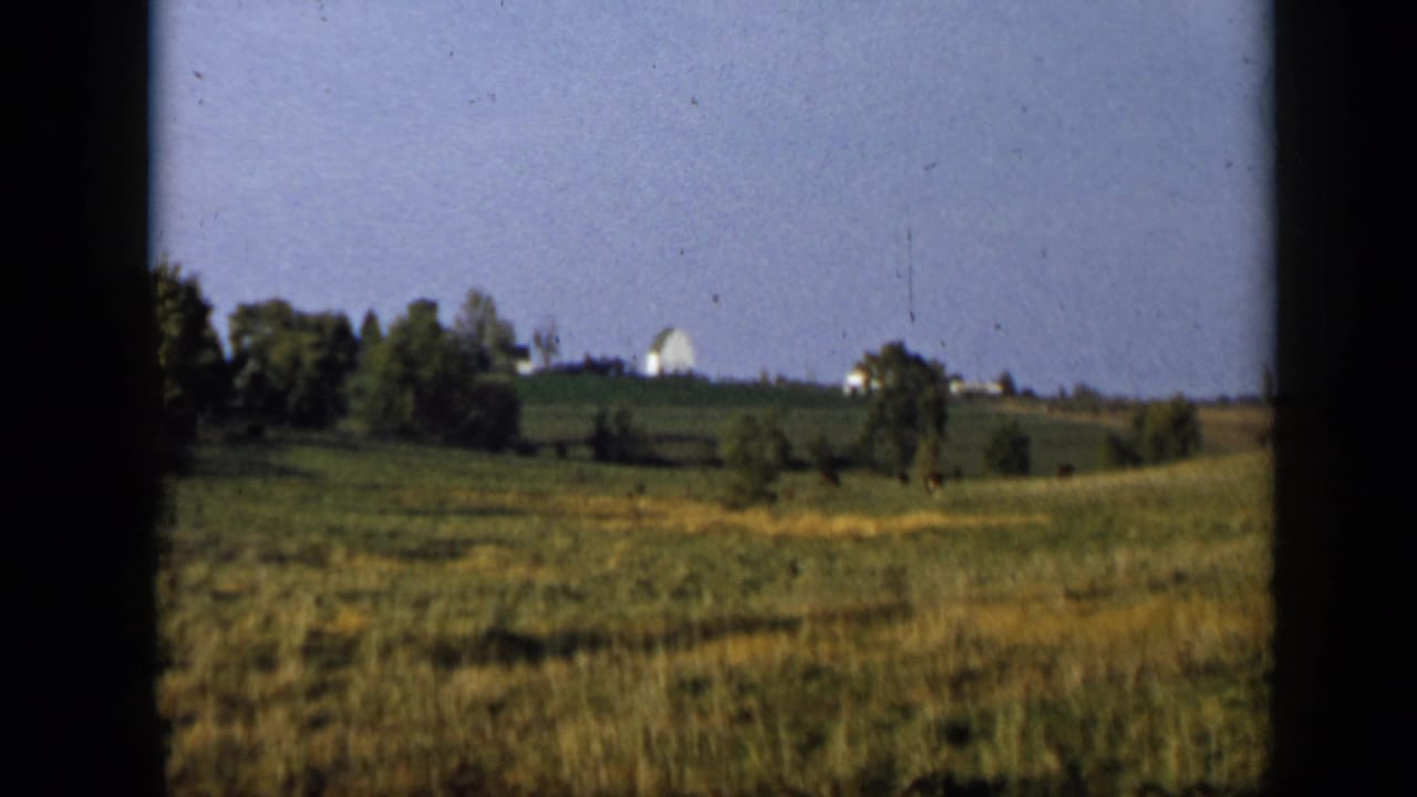 Aledo, Texas home-movie view of rolling green fields, scattered trees, and distant light-colored buildings along the horizon.
