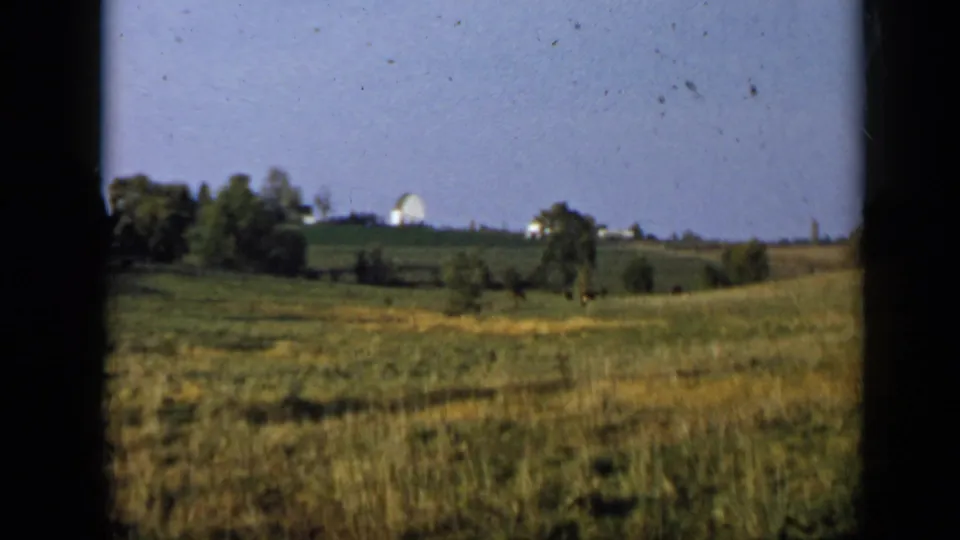 1959 8mm frame from Aledo showing pasture-like grass, trees, and a distant white structure beyond the field.