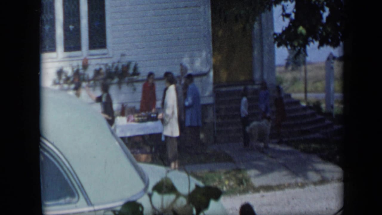 Women gather near a table outside a white house with a parked car in the foreground.