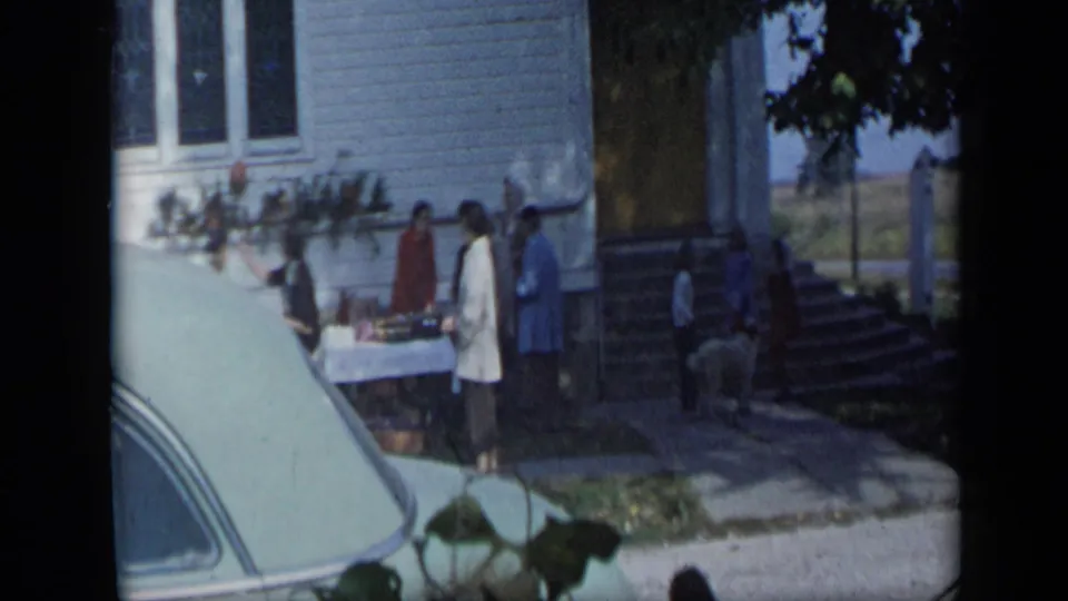 Aledo, Texas 1959 home-movie frame showing women around an outdoor sale table beside a white clapboard house and parked car.