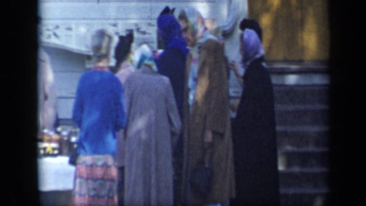 Older women in colorful coats and headscarves gather beside a table of goods outside the Aledo house in 1959 footage.