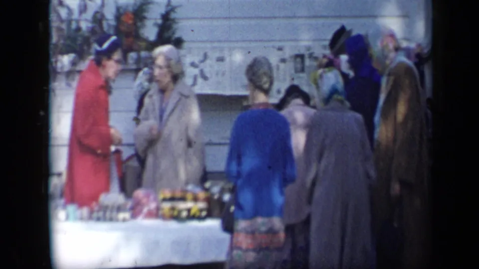 A 1959 Aledo outdoor sale frame shows a red-coated woman talking with an older woman near jars or small goods on a table.