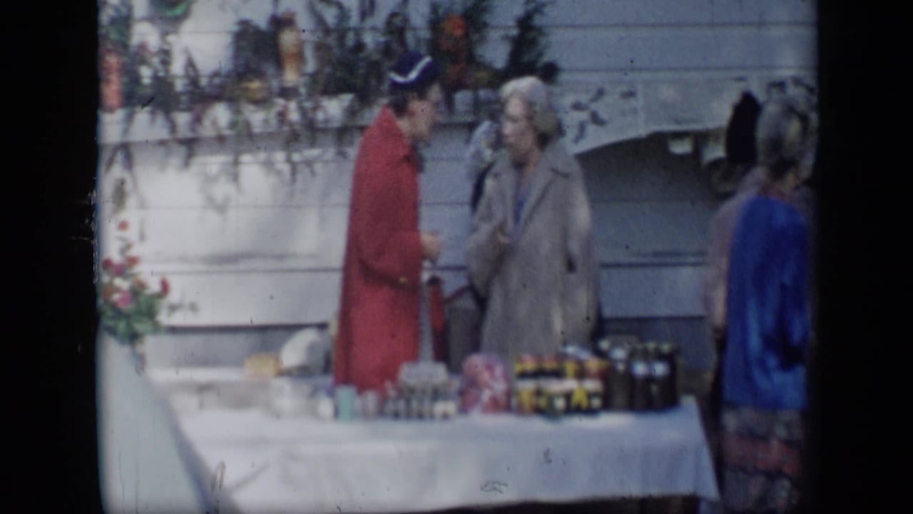 People stand beside a white-clothed outdoor table with jars and food items.
