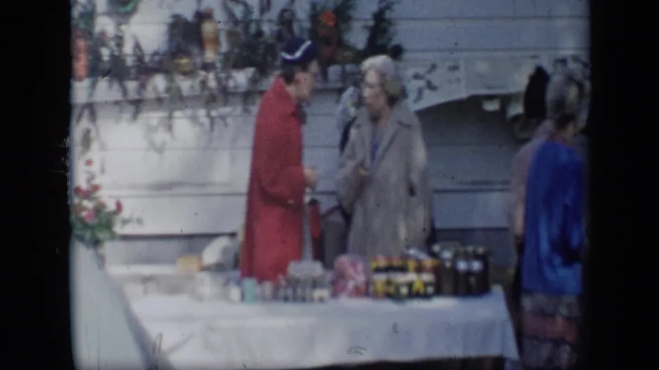 In 1959 Aledo, Texas, a person in a red jacket speaks with an older woman beside a white table of jars and food items.