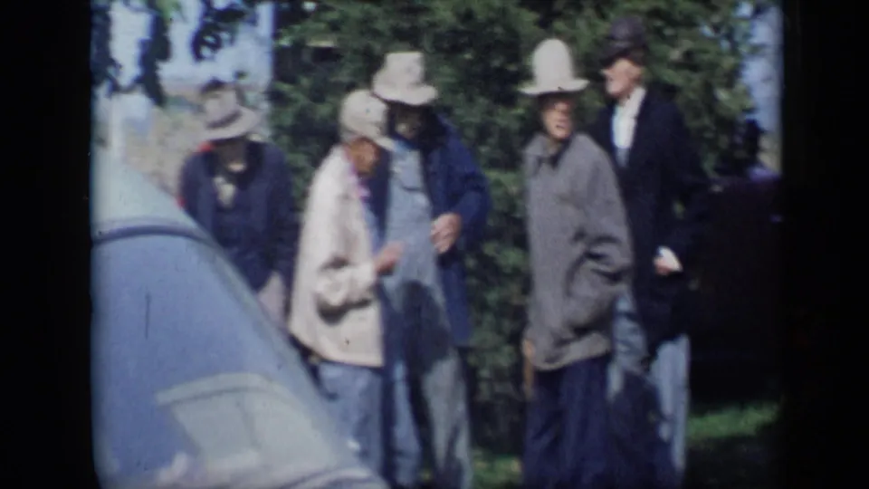 A candid 1959 home-movie view shows adults in hats and coats mingling beside a light-colored car and leafy background in Aledo, Texas.
