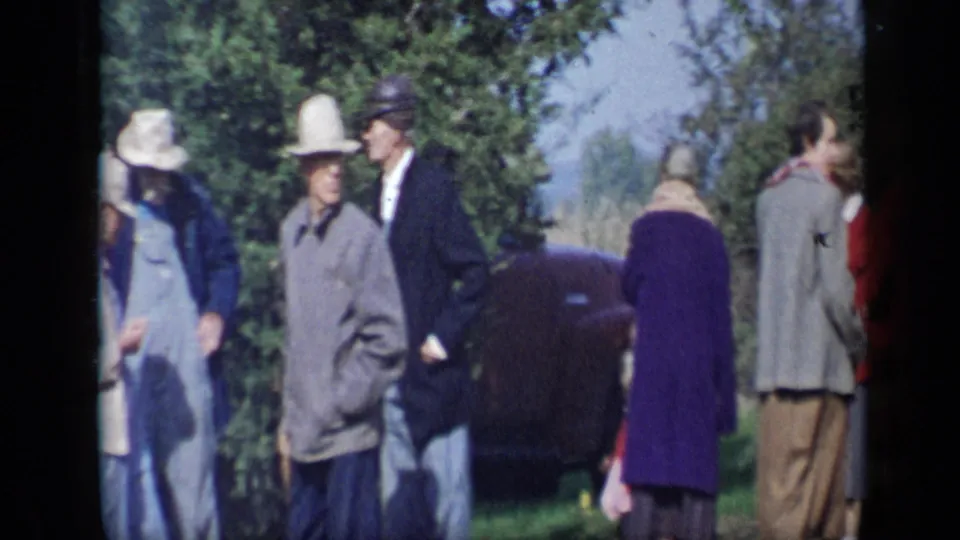 At the end of the Aledo, Texas, clip, people in coats and hats move past a dark car under trees in a casual outdoor gathering.