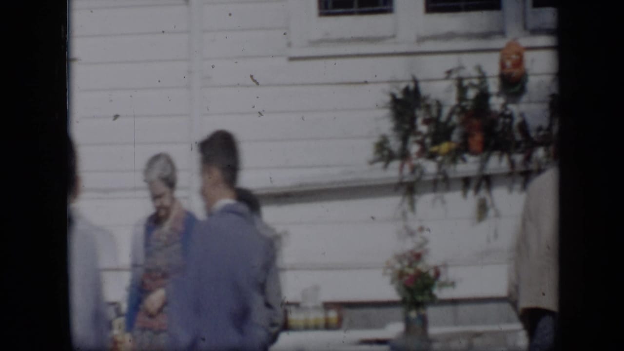 In Aledo, Texas, a 1959 home-movie frame shows adults near vases and greenery beside a white clapboard wall.