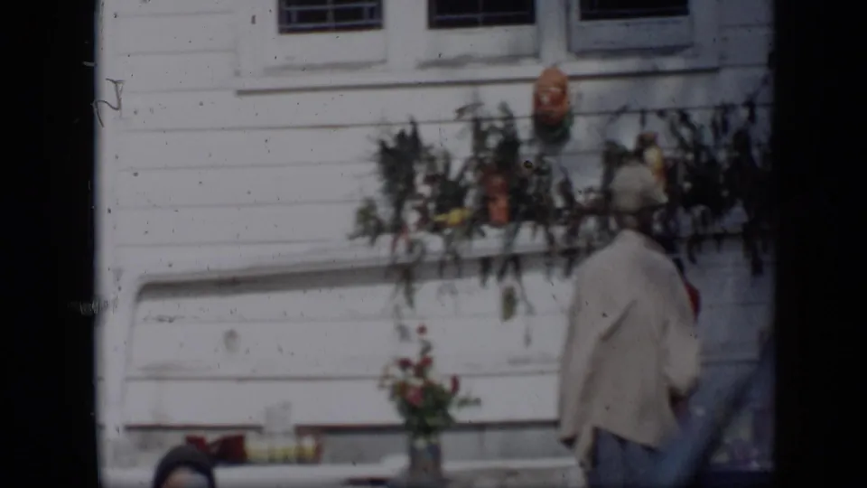 A 1959 Aledo frame shows a woman in a light coat and hat passing flowers and hanging decorations on a clapboard exterior.