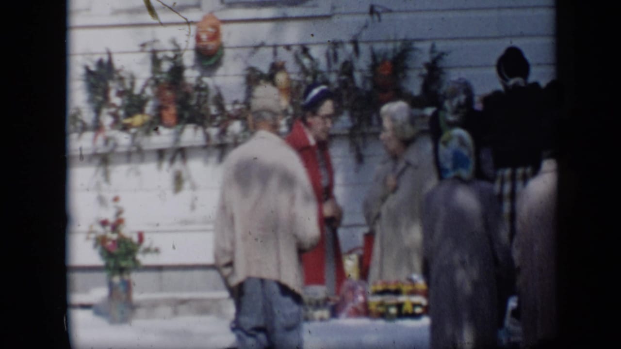 Adults stand near a white-covered produce table beside a decorated clapboard wall.