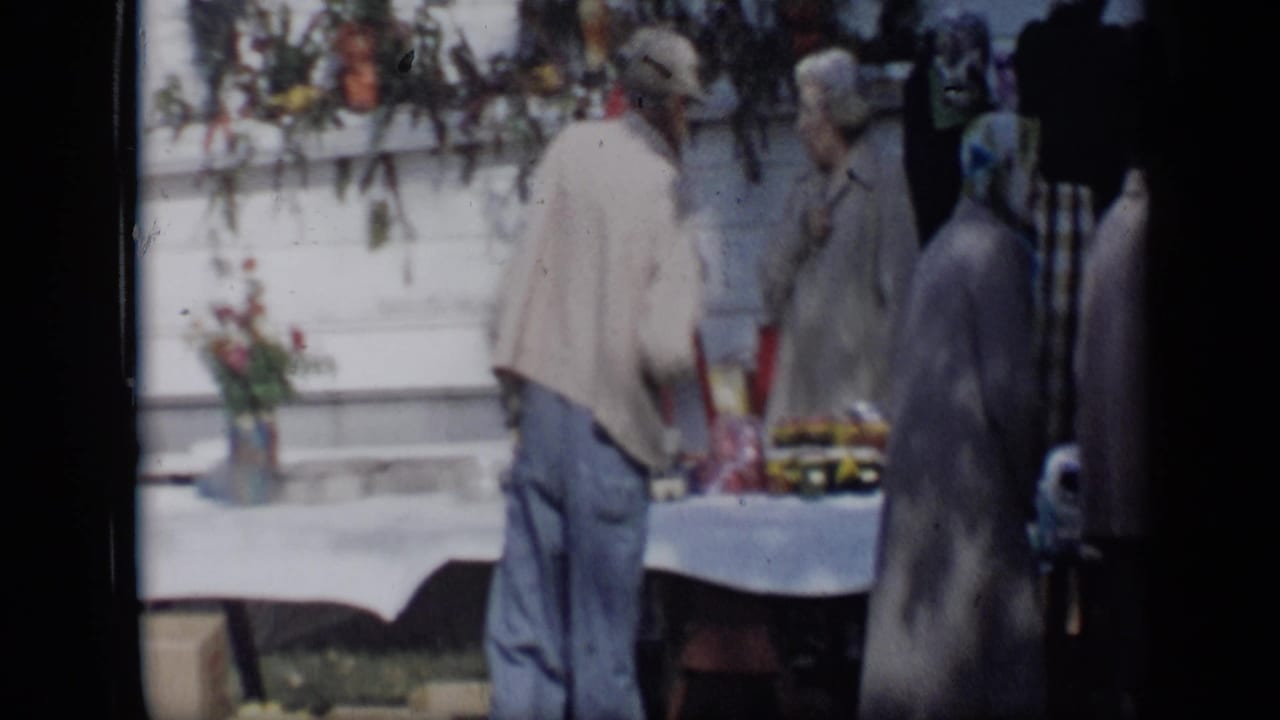1959 Aledo, Texas frame with a man in a light jacket at an outdoor produce table as women stand and talk beside him.
