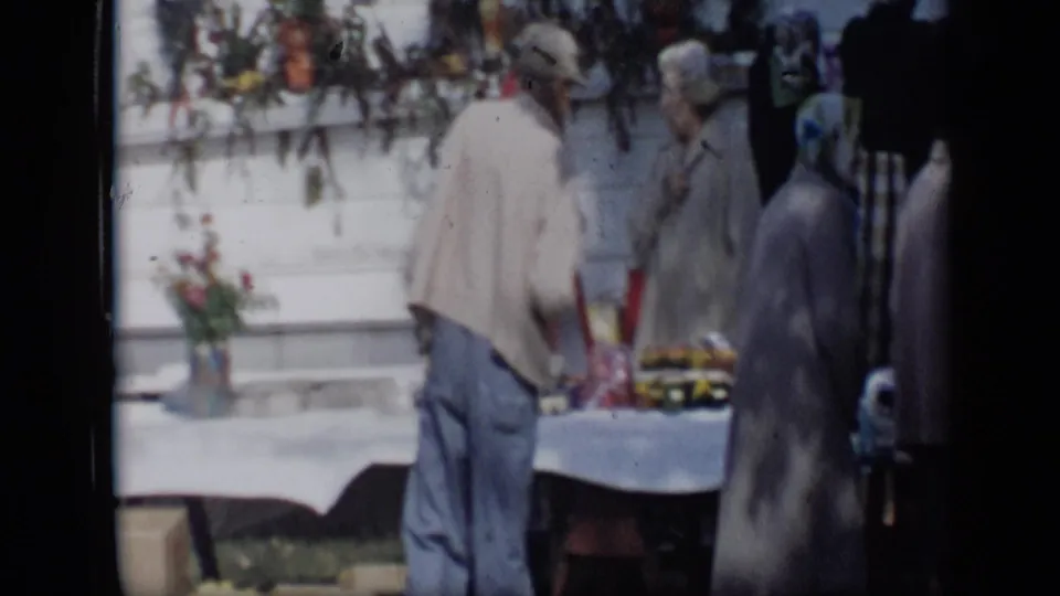 1959 Aledo, Texas frame with a man in a light jacket at an outdoor produce table as women stand and talk beside him.