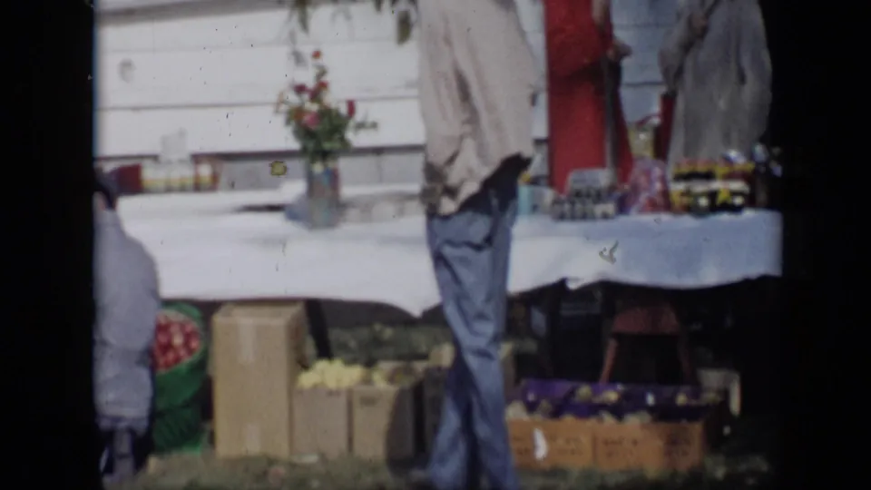1959 Aledo, Texas home-movie frame showing the man turned near a white-covered produce table with boxes and baskets below.