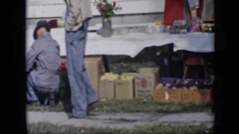 1959 Aledo, Texas frame with a seated person, a standing man, produce boxes, baskets, and a white-covered table at an outdoor stand.