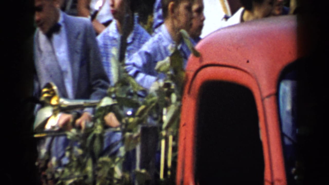 In 1959 Aledo, Texas footage, a boy in a light suit and bow tie holds a trumpet beside a red vehicle as people gather nearby.