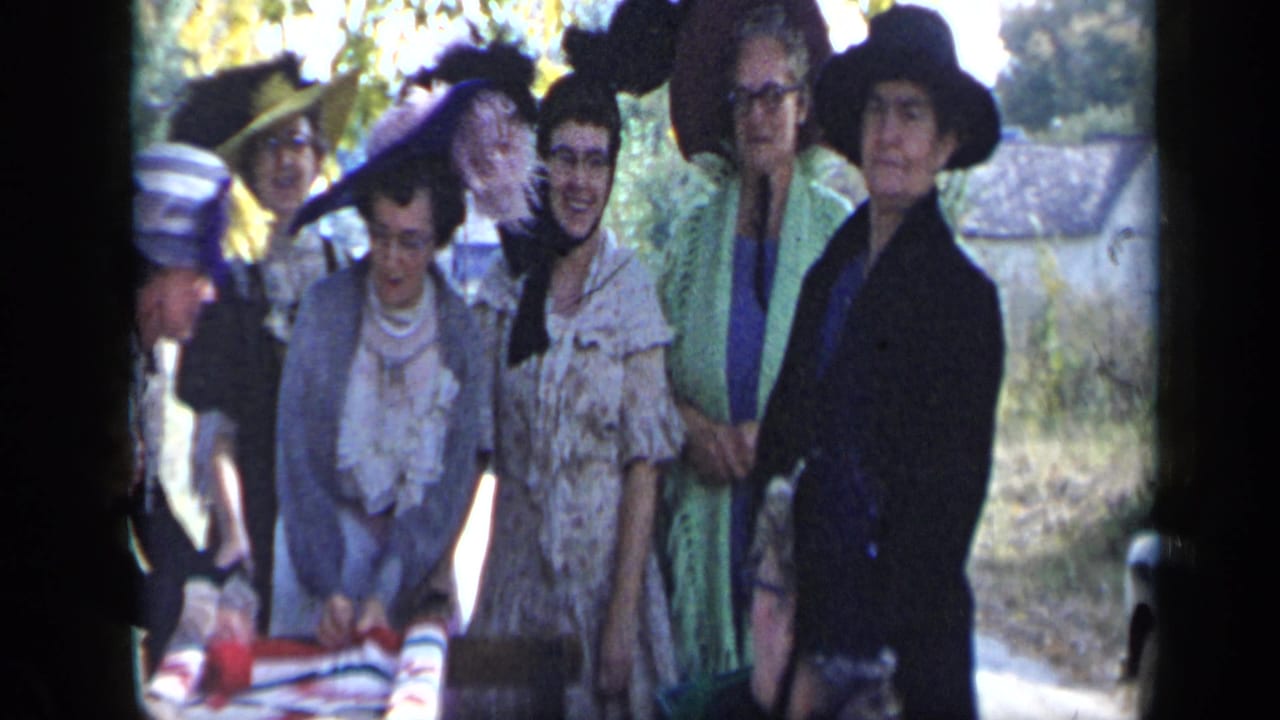 Group of women in wide hats and dresses gather around an outdoor table.