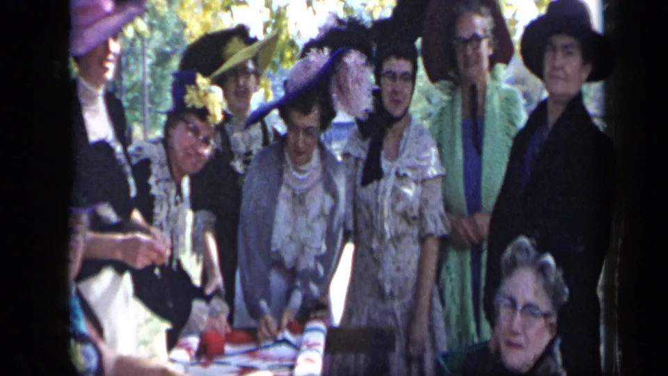 Several women in hats and dresses lean toward the camera near a covered table in the Aledo, Texas celebration footage.