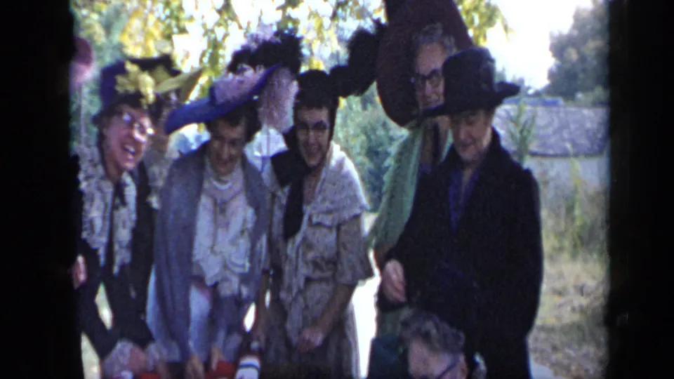 Women in dark and light hats stand together near the table, with trees and a roadside or open outdoor area behind them.