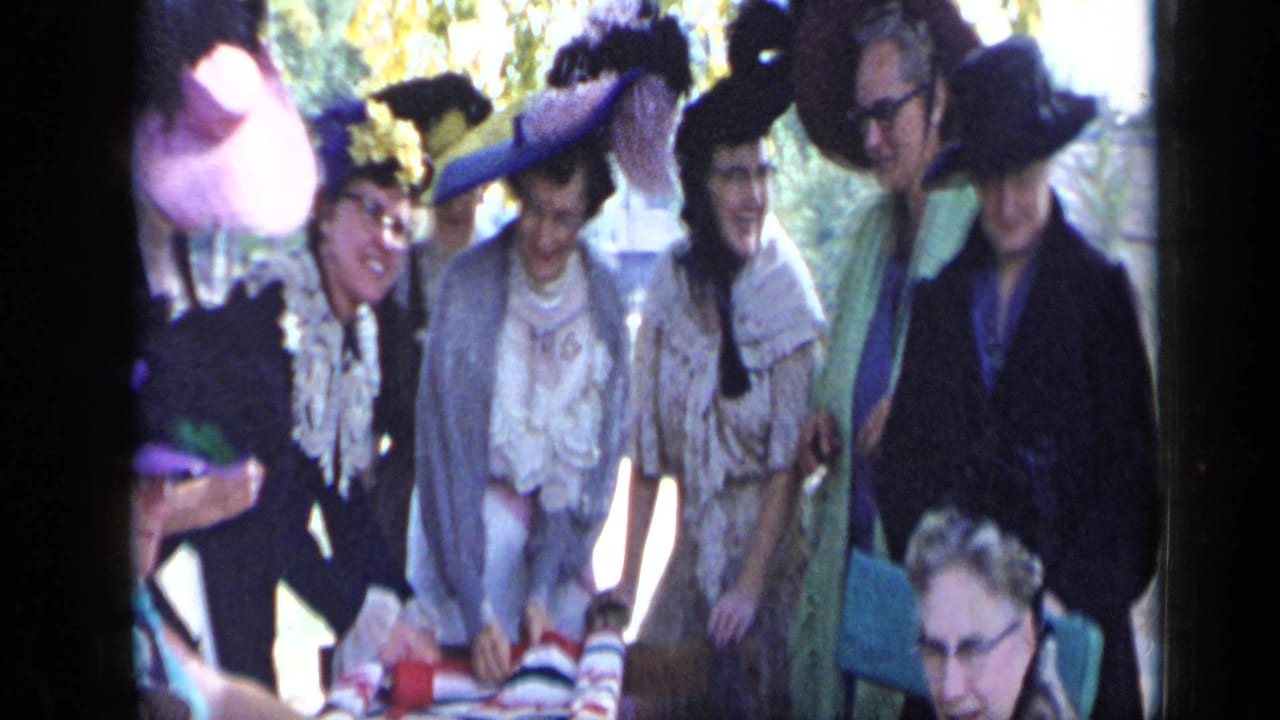 In Aledo, Texas in 1959, women wearing elaborate feathered hats stand around an outdoor table in dappled sunlight.