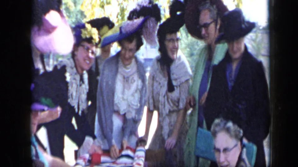 In Aledo, Texas in 1959, women wearing elaborate feathered hats stand around an outdoor table in dappled sunlight.