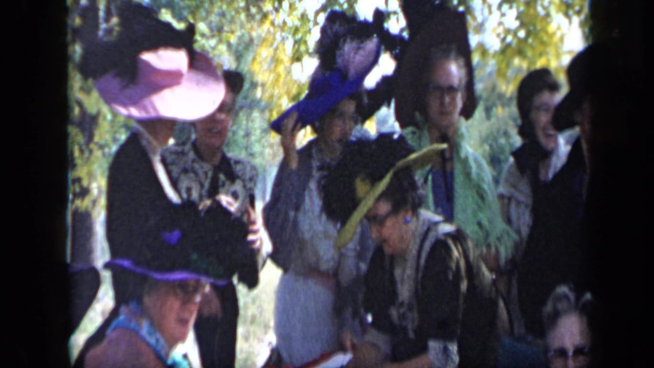 Women in wide-brimmed and feathered hats converse outdoors near a table.