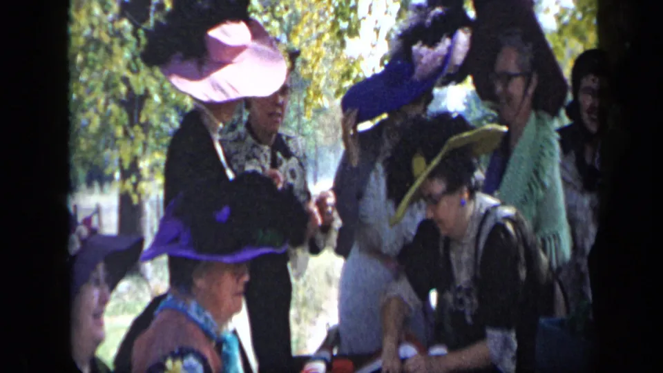 A 1959 Aledo, Texas home-movie frame shows women in ornate hats leaning and talking near a table under trees.
