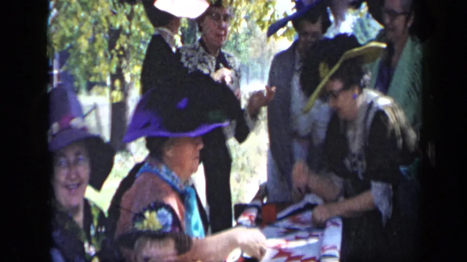 Women in Aledo, Texas in 1959 gather beside a red-and-white covered table, with feathered hats and trees visible around them.