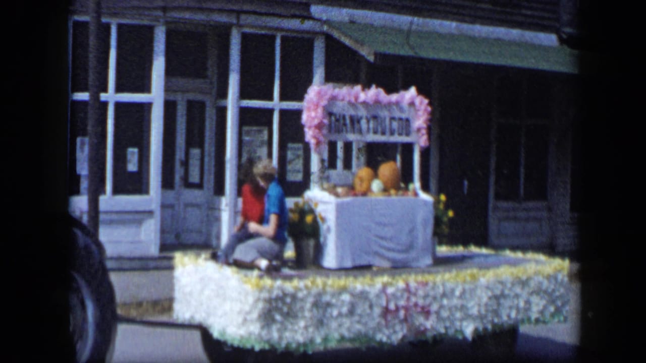 Decorated parade float with pumpkins and a THANK YOU GOD sign passing storefronts.