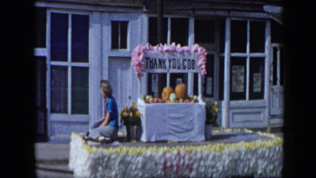 Woman seated on a flower-trimmed parade float in Aledo, Texas, with pumpkins, flowers, and a THANK YOU GOD sign in 1959 footage.
