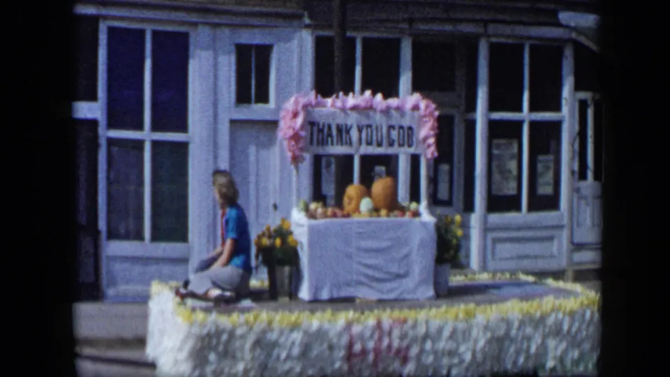 Woman seated on a flower-trimmed parade float in Aledo, Texas, with pumpkins, flowers, and a THANK YOU GOD sign in 1959 footage.