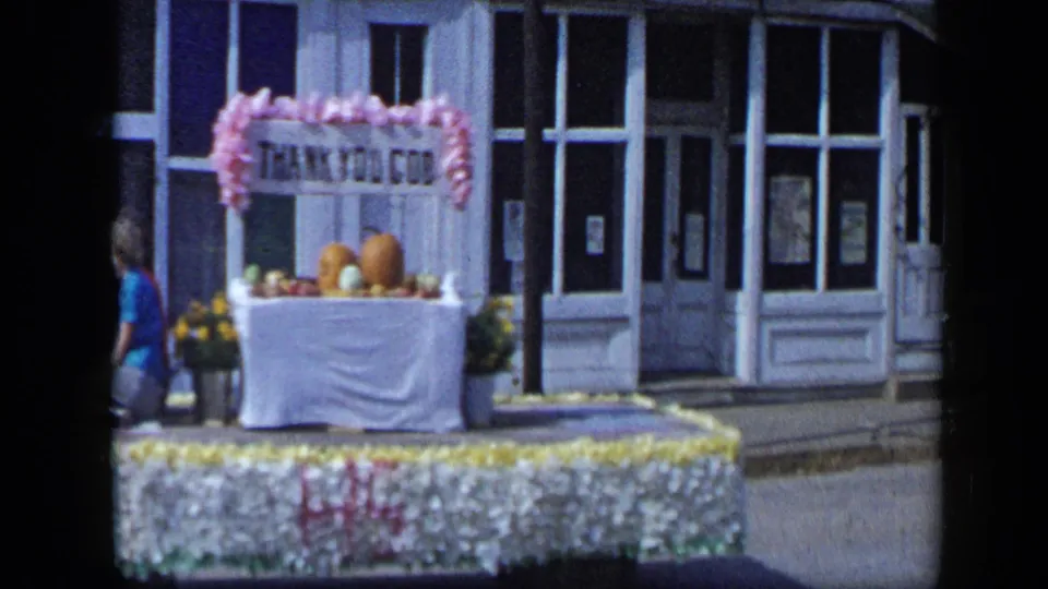 Aledo, Texas parade float in 1959 with pink-fringed THANK YOU GOD sign, pumpkins, flowers, and storefronts behind it.