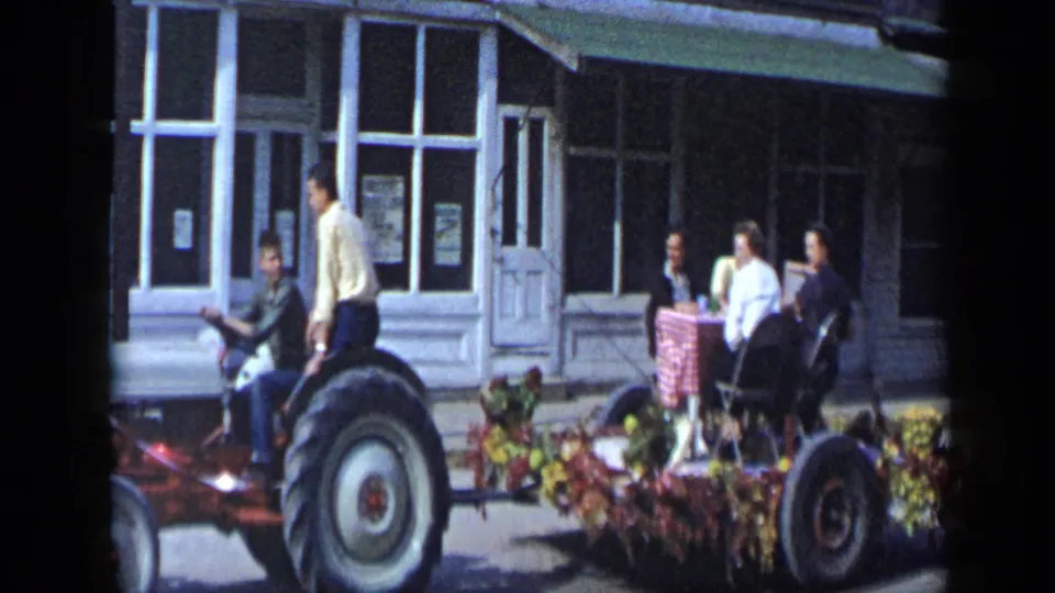 Tractor pulling a decorated parade float past storefronts in 1959 Aledo, Texas, with riders and flower trim visible.