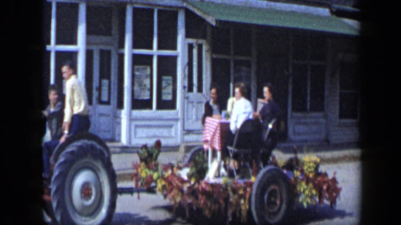 Tractor-pulled parade float with women gathered around a table in front of storefront windows.